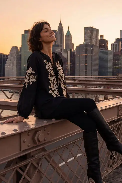 Blouse noire à broderies florales blanches, portée avec un pantalon et des bottes noires sur le pont de Brooklyn, avec la skyline de New York